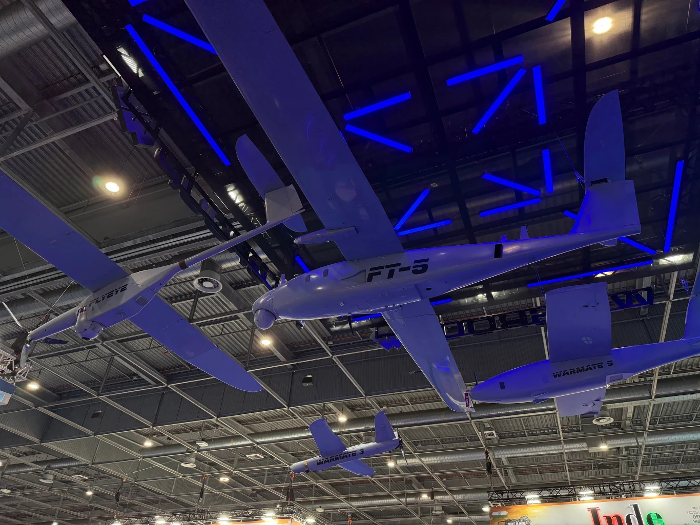 Warmate, FlyEye, FT5 Łoś na targach Eurosatory 2026. Kilka bezzałogowych samolotów prezentacyjnych zawieszonych pod sufitem nowoczesnej hali wystawowej, oświetlonych niebieskimi światłami LED.