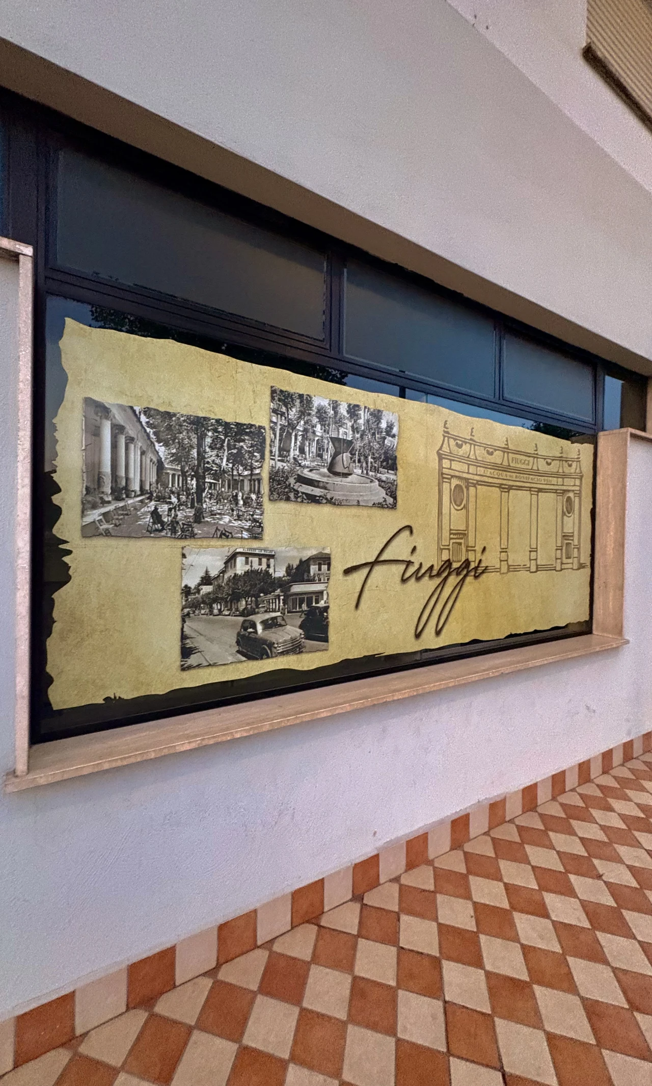 Tablica z historycznymi czarno-białymi fotografiami oraz szkicem budynku, ozdobiona napisem „Fiuggi”, umieszczona na tle jasnej ściany nad mozaikową posadzką.