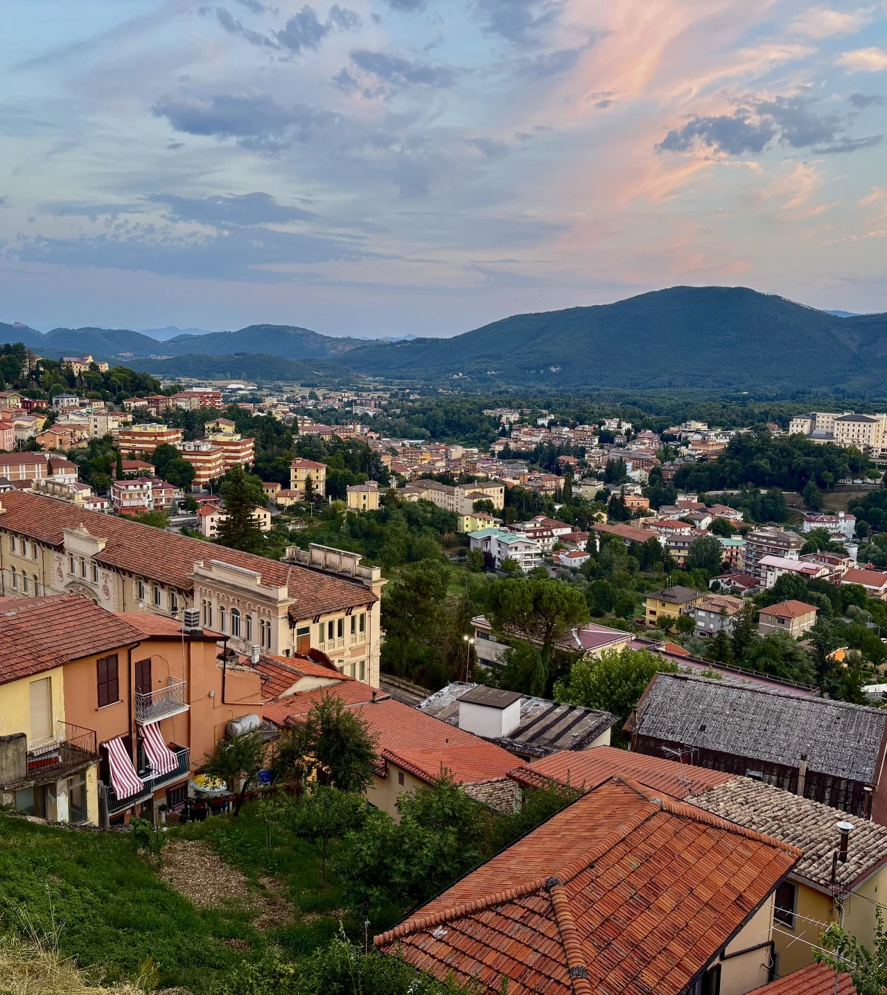 Panorama miasta z licznymi budynkami o czerwonych dachach otoczonymi zielenią, w tle rozciągające się wzgórza pod miękkim, pastelowym niebem.