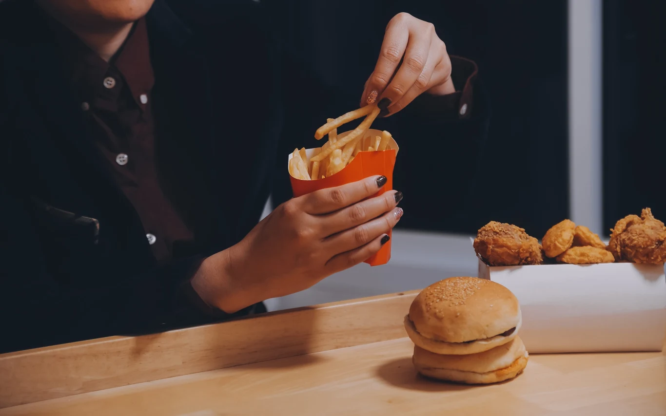 Ręka sięga po frytki z czerwonego opakowania, obok na tacy znajduje się hamburger oraz kawałki panierowanego kurczaka.