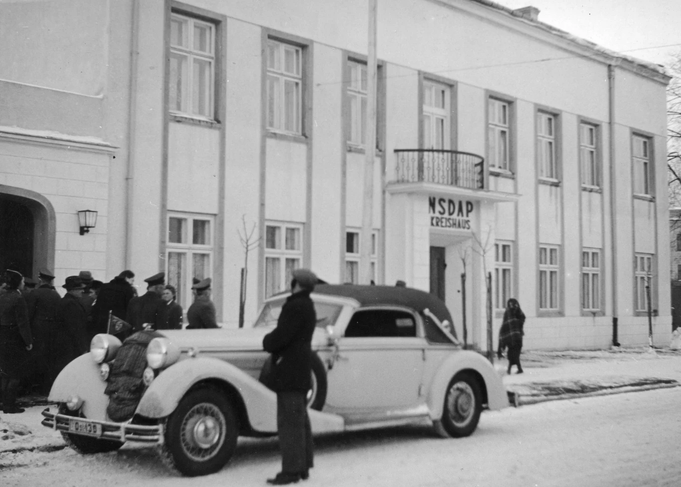 Elegancki samochód zaparkowany na ośnieżonej ulicy przed budynkiem z szyldem NSDAP Kreishaus, na chodniku kilka osób w płaszczach i kapeluszach, jedna z nich stoi przy samochodzie.