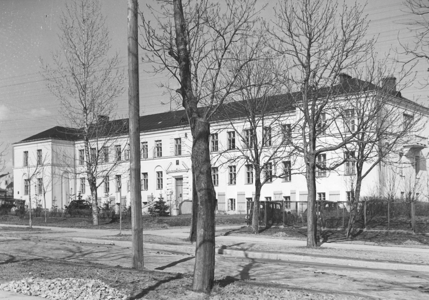 Duży, dwukondygnacyjny budynek o jasnej elewacji z licznymi oknami, otoczony drzewami bez liści, z przodu szeroka, pusta ulica i chodnik.
