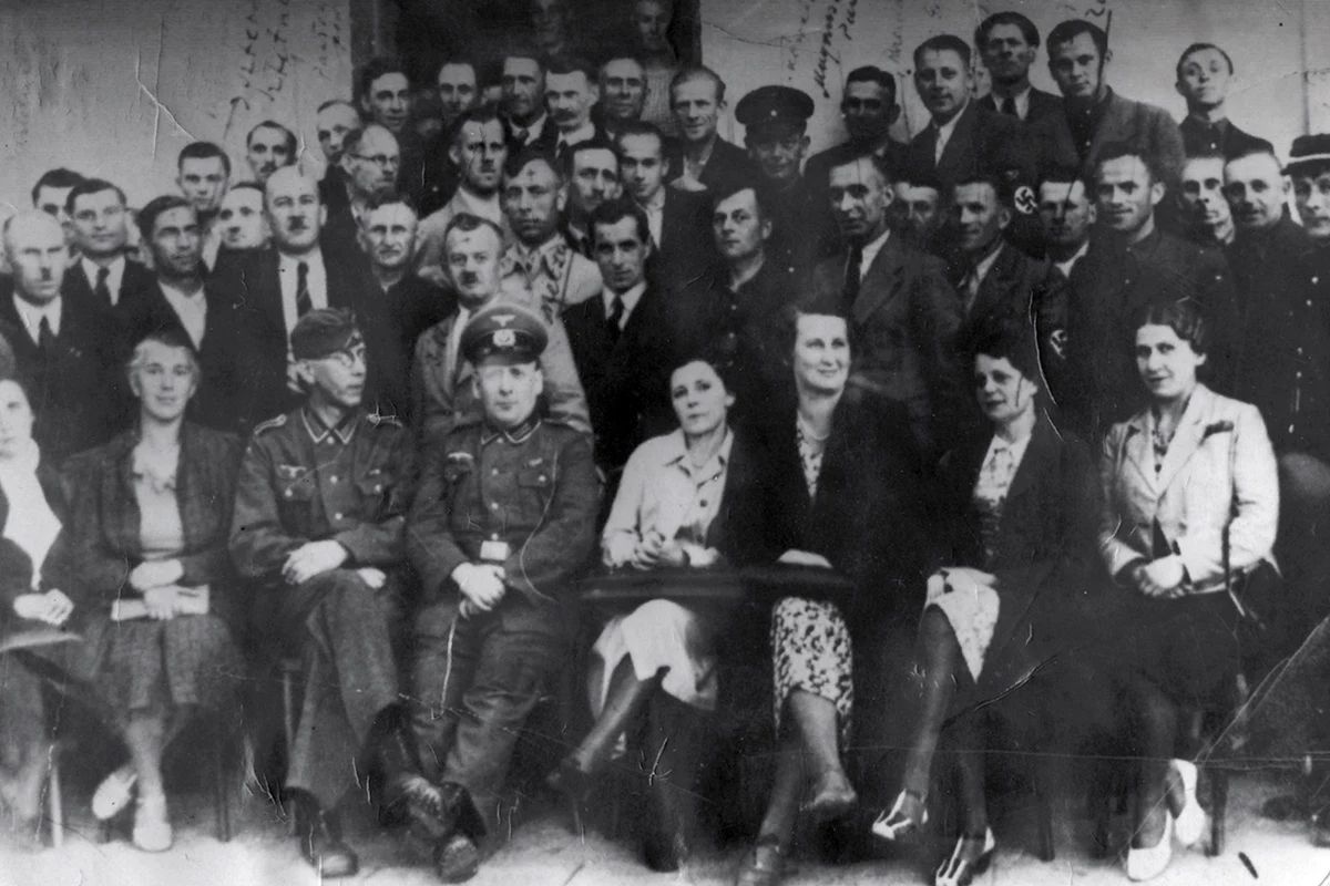 Grupa kilkudziesięciu dorosłych osób w formalnych strojach, z przodu siedzi kilku mężczyzn w mundurach wojskowych oraz kobiety w sukienkach, pozostali stoją w kilku rzędach z poważnymi minami, całość sfotografowana w czerni i bieli.