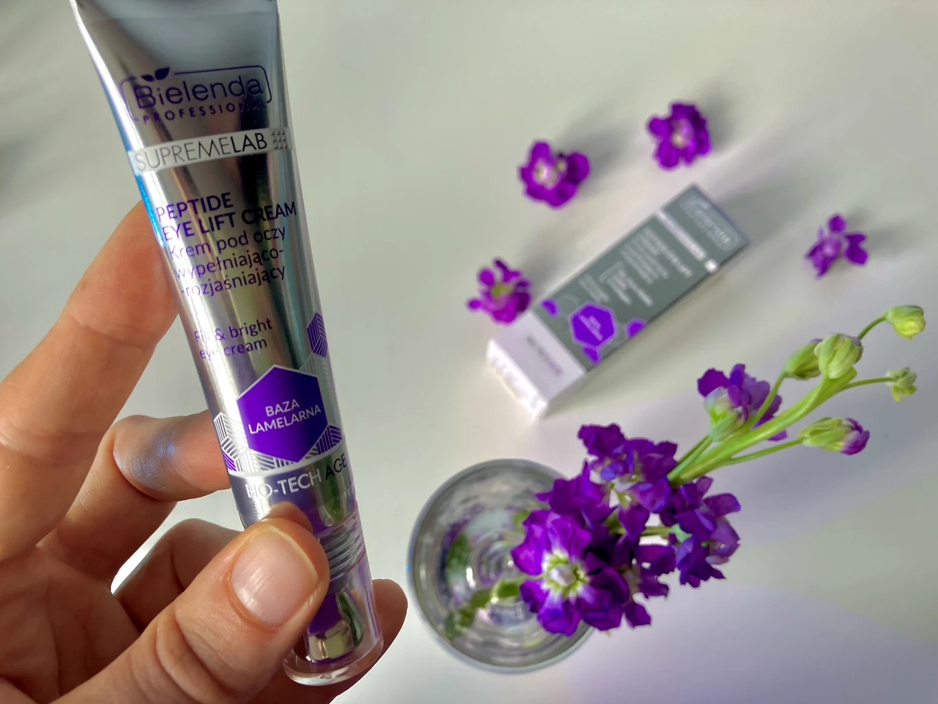 Krem pod oczy pomaga wygładzać drobne zmarszczki mimiczne Tuba kremu pod oczy Bielenda SupremeLab Peptide Lift Cream w dłoni, obok szklanka z fioletowymi kwiatami oraz kartonik z opakowania kremu na jasnym tle.