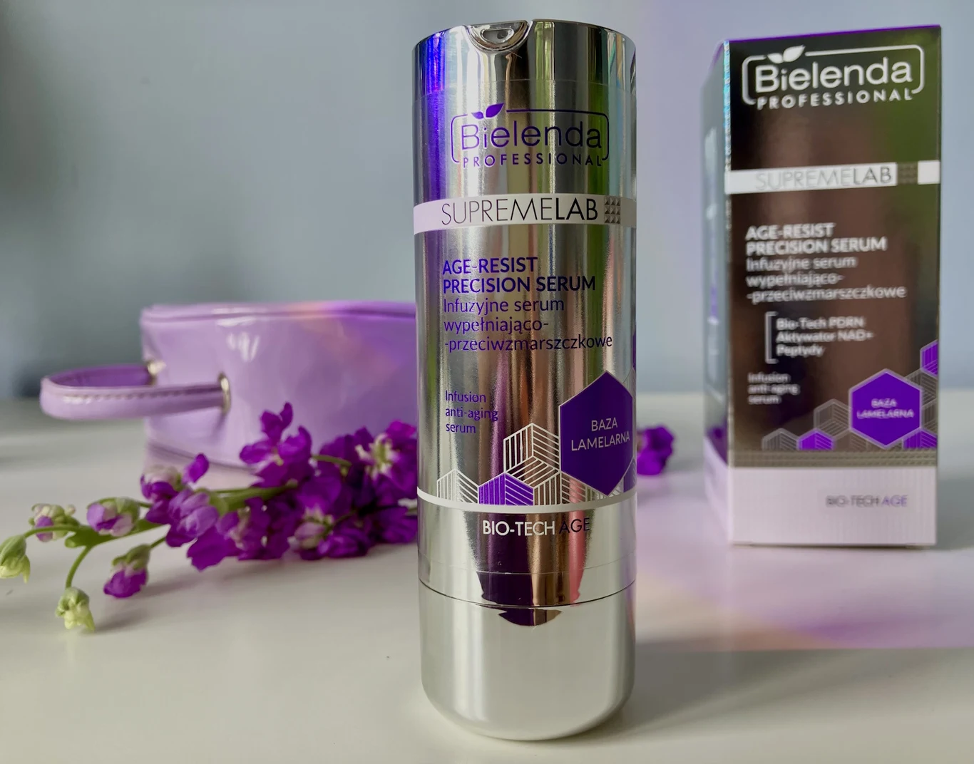 Serum daje efekt natychmiastowego napięcia skóry Butelka serum Bielenda SupremeLab Age Resist Precision Serum z fioletowymi akcentami ustawiona na jasnej powierzchni, obok opakowania produktu i dekoracyjnych kwiatów w odcieniach fioletu.