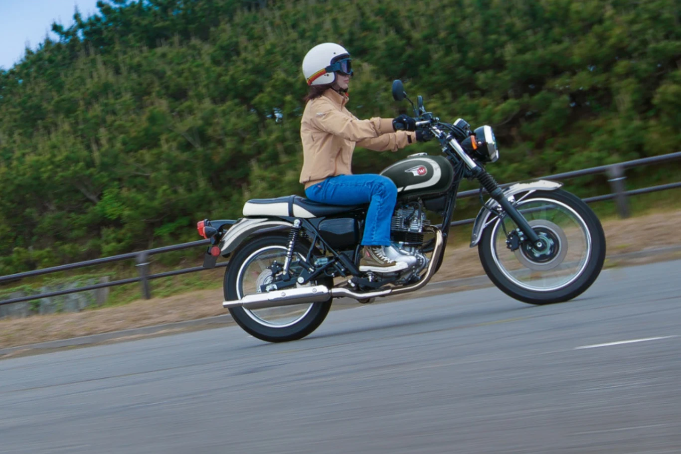 Osoba w kasku i kurtce jedzie klasycznym motocyklem po asfaltowej drodze, w tle widoczne drzewa iglaste.