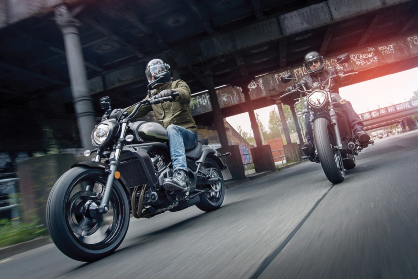 Dwóch motocyklistów w kaskach i kurtkach jedzie miejską ulicą pod stalowym wiaduktem, prowadząc sportowe motocykle w dynamicznej pozie.