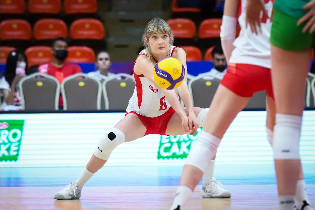 Zawodniczka w biało-czerwonym stroju skutecznie odbiera piłkę do siatkówki podczas meczu, skupiona i przygotowana do kolejnego ruchu, w otoczeniu innych zawodniczek na boisku.