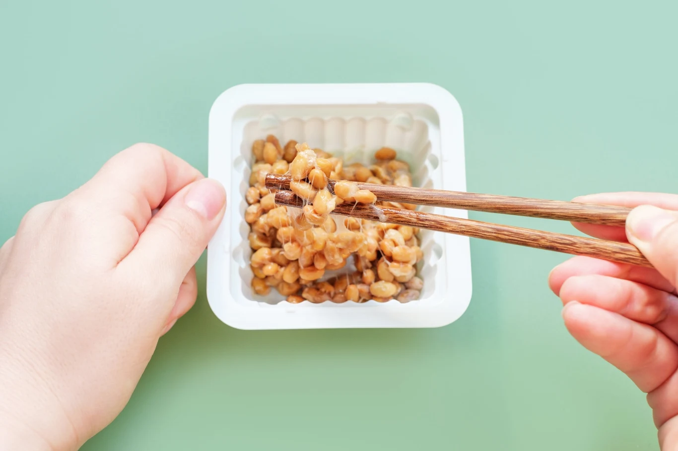 W takim opakowaniu najczęściej można zakupić natto Porcja fermentowanej soi natto w plastikowym pojemniku, podnoszona pałeczkami przez dwie ręce na tle jasnozielonej powierzchni.
