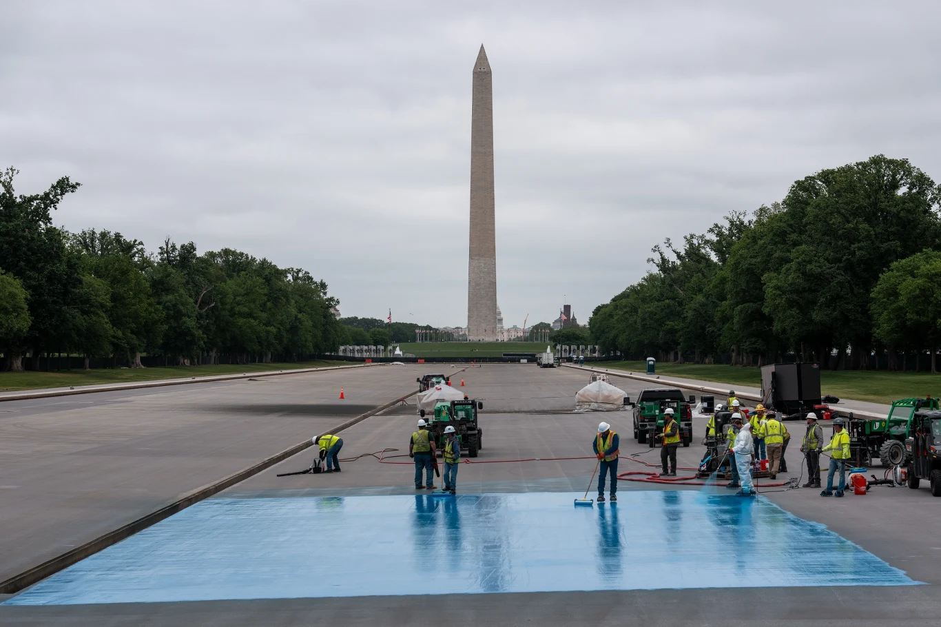 Robotnicy w kamizelkach odblaskowych rozkładają jasnoniebieską folię na dużej, płaskiej powierzchni w otoczeniu maszyn budowlanych, na pierwszym planie, z Washington Monument w tle pośrodku alei otoczonej drzewami.