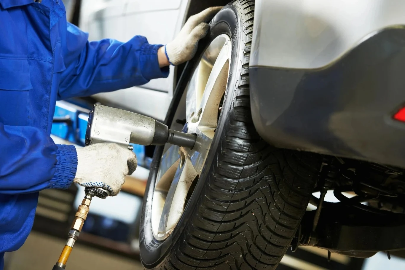 Mechanik w niebieskim kombinezonie wykorzystuje klucz pneumatyczny do odkręcania śrub w kole samochodu unoszonego na podnośniku.