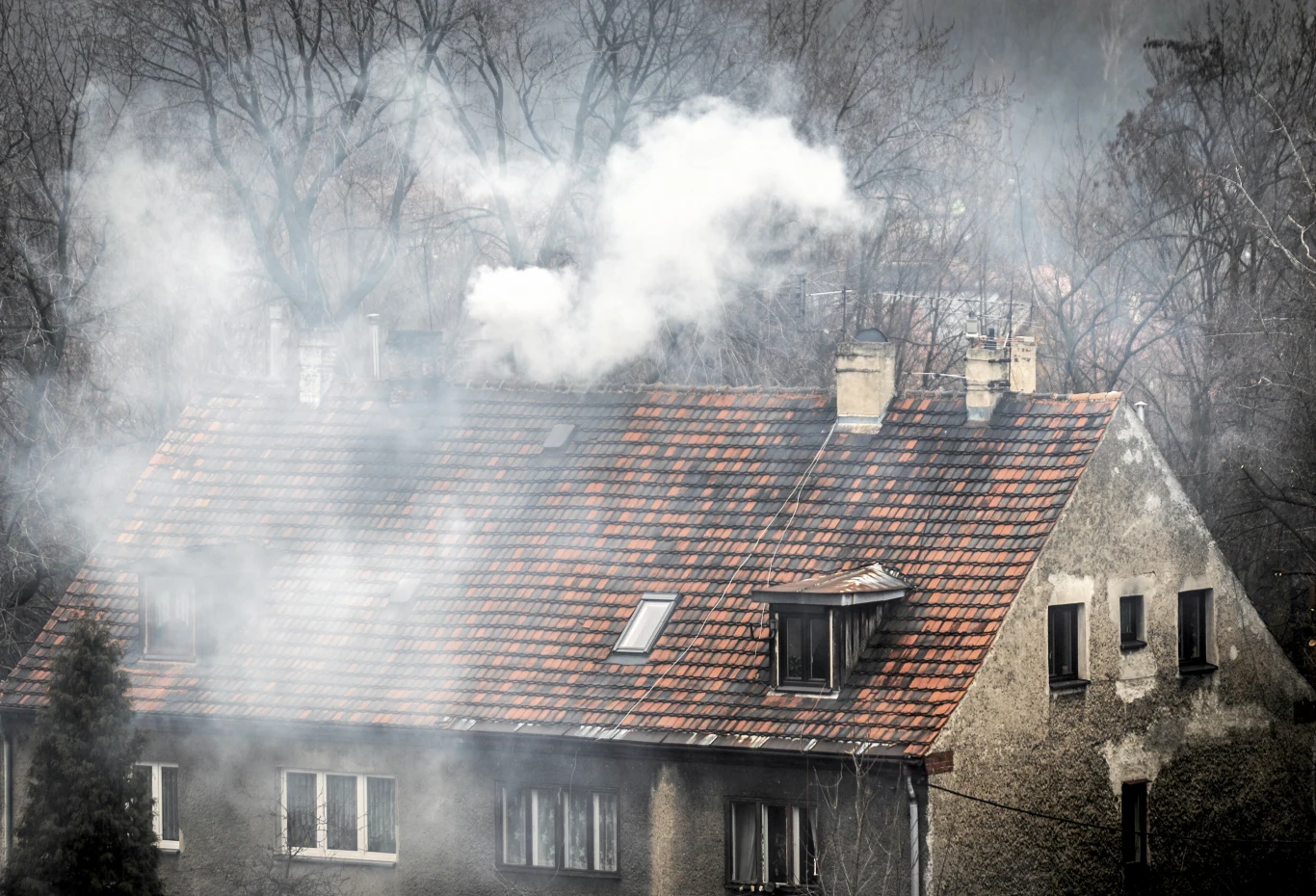 Dane wskazują, że w najbardziej zanieczyszczonych miastach w Polsce jest coraz więcej smogu Stary budynek mieszkalny z czerwonym dachem, z którego kominów wydobywa się gęsty dym, otoczony bezlistnymi drzewami.