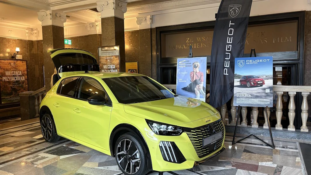 Jaskrawożółty samochód Peugeot z otwartą klapą bagażnika zaprezentowany w eleganckim wnętrzu, obok stoją bannery promocyjne marki i plakat reklamowy.