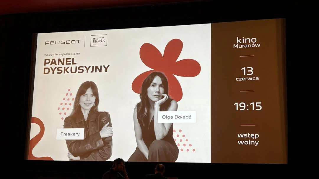 Duży ekran z zapowiedzią panelu dyskusyjnego, na którym widoczne są zdjęcia dwóch kobiet – Freakery i Olgi Bołądź, wraz z informacją o dacie, godzinie oraz miejscu wydarzenia w kinie Muranów; po prawej stronie wyraźne napisy i grafika czerwonego kwiatu.