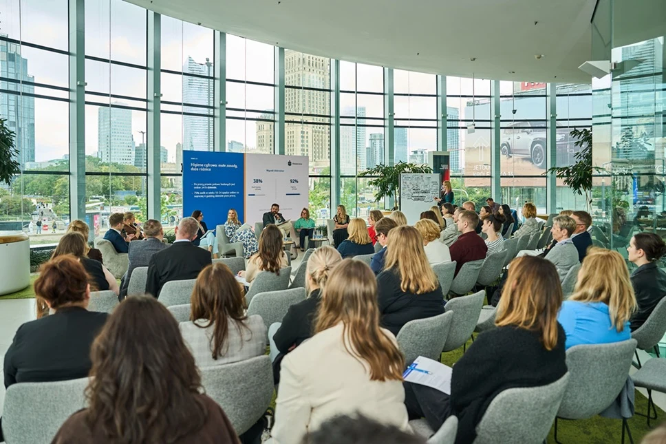 Grupa uczestników siedzi na wygodnych krzesłach w przeszklonej sali konferencyjnej, słuchając panelu dyskusyjnego z udziałem kilku prelegentów rozmawiających przy stole na tle dużego niebieskiego baneru informacyjnego.