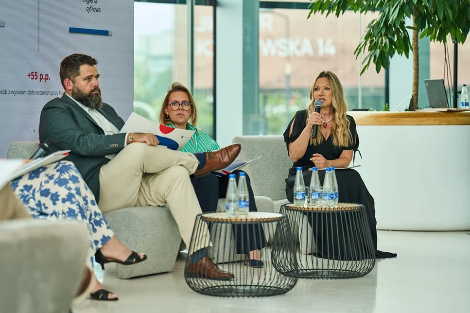 Trzy osoby siedzące na nowoczesnych fotelach podczas panelu dyskusyjnego, jedna kobieta trzyma mikrofon i mówi, na stolikach stoją butelki z wodą mineralną.
