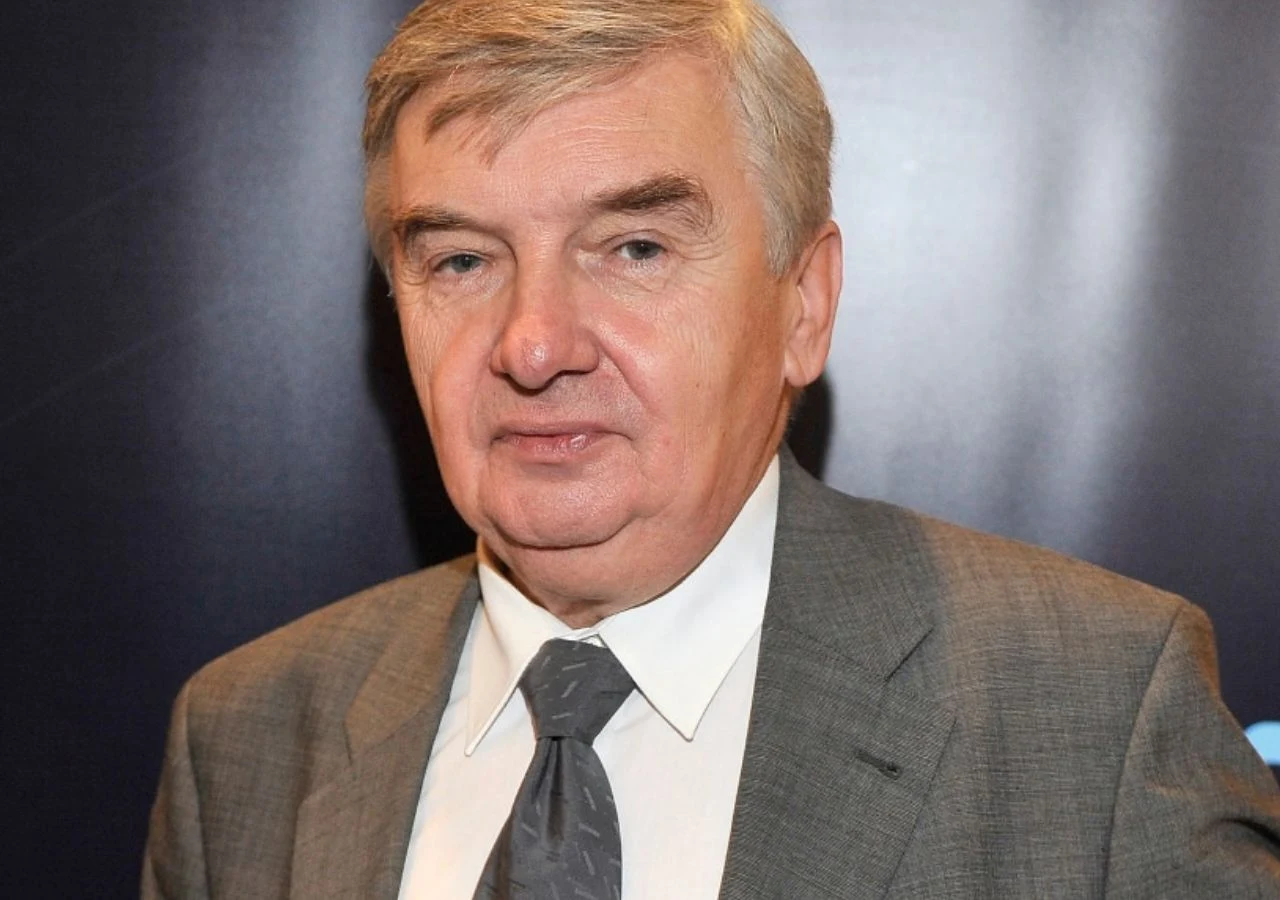 Tadeusz Sznuk Starszy mężczyzna w szarym garniturze, białej koszuli i szarej krawacie na ciemnym jednolitym tle.