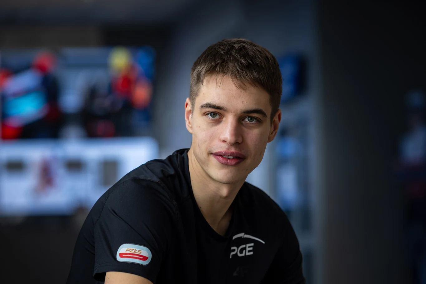 Młody mężczyzna z krótkimi, brązowymi włosami i niebieskimi oczami, ubrany w czarną koszulkę sportową z widocznymi logotypami PGE oraz PZM, patrzy lekko uśmiechnięty w stronę obiektywu.