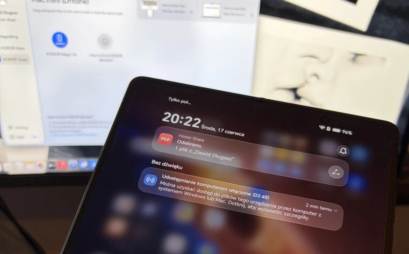 Ekran smartfona z widocznymi powiadomieniami o nowej wiadomości i udostępnianiu komputera, na tle monitora komputerowego wyświetlającego ustawienia systemowe i fragment wydruku graficznego.