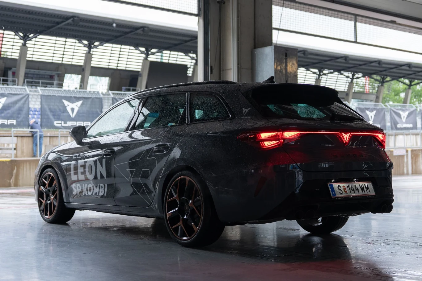 Czarny samochód Cupra Leon Sportstourer z przyciemnianymi szybami i charakterystycznymi tylnymi światłami LED zaparkowany w zadaszonym miejscu przy torze wyścigowym.