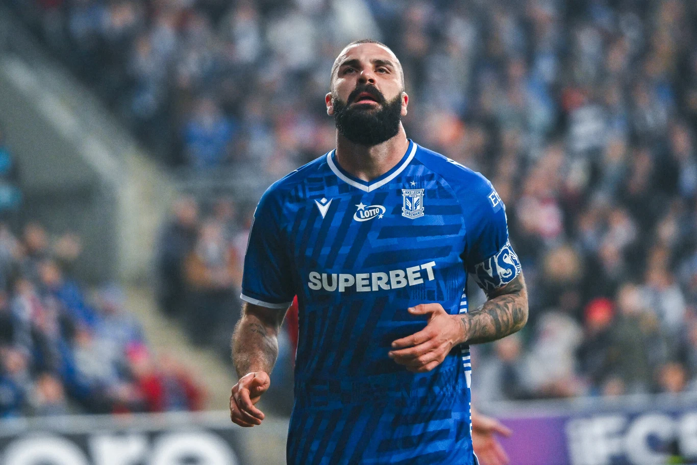 Piłkarz w niebieskiej koszulce Lecha Poznań z widocznymi sponsorami na tle rozmytego stadionu i kibiców.