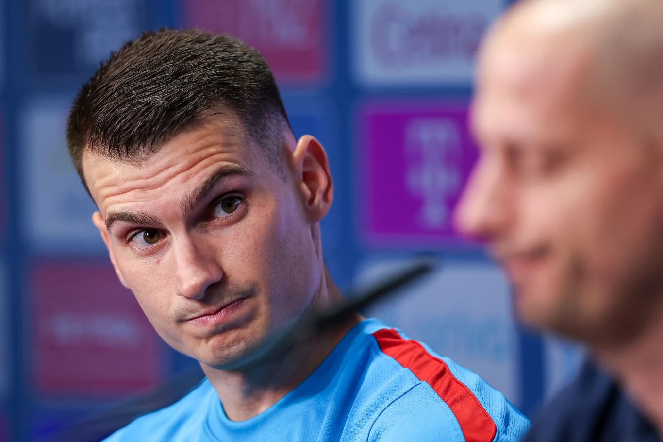 Dwóch mężczyzn podczas konferencji prasowej, jeden z nich w sportowej koszulce z czerwonymi wstawkami, drugi nieco rozmyty na pierwszym planie, tło z rozmytymi logotypami sponsorów.