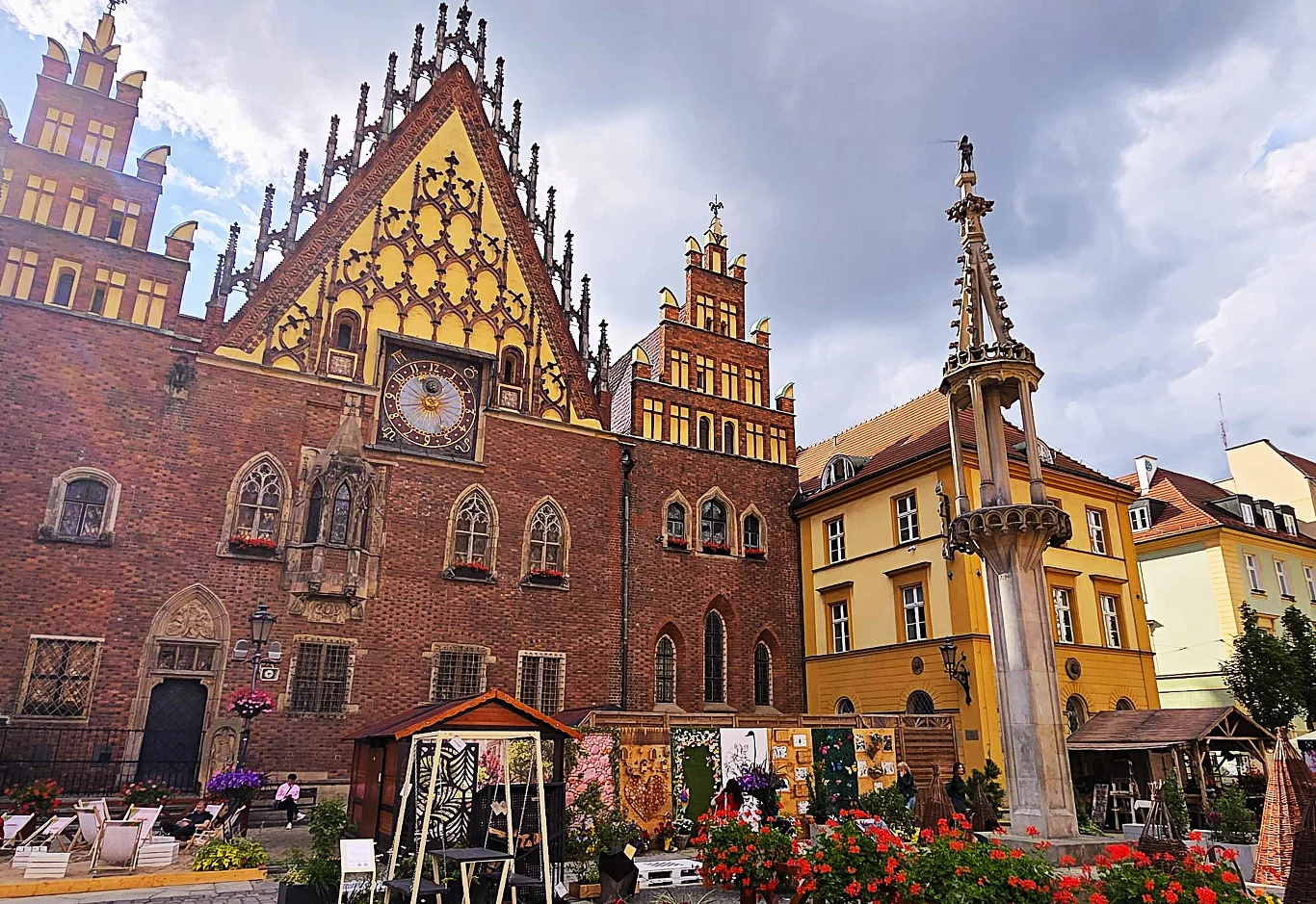 Gotycki ratusz z bogato zdobioną fasadą i zegarem astronomicznym, obok kamienna fontanna otoczona kwiatami oraz kolorowe kamienice w tle.