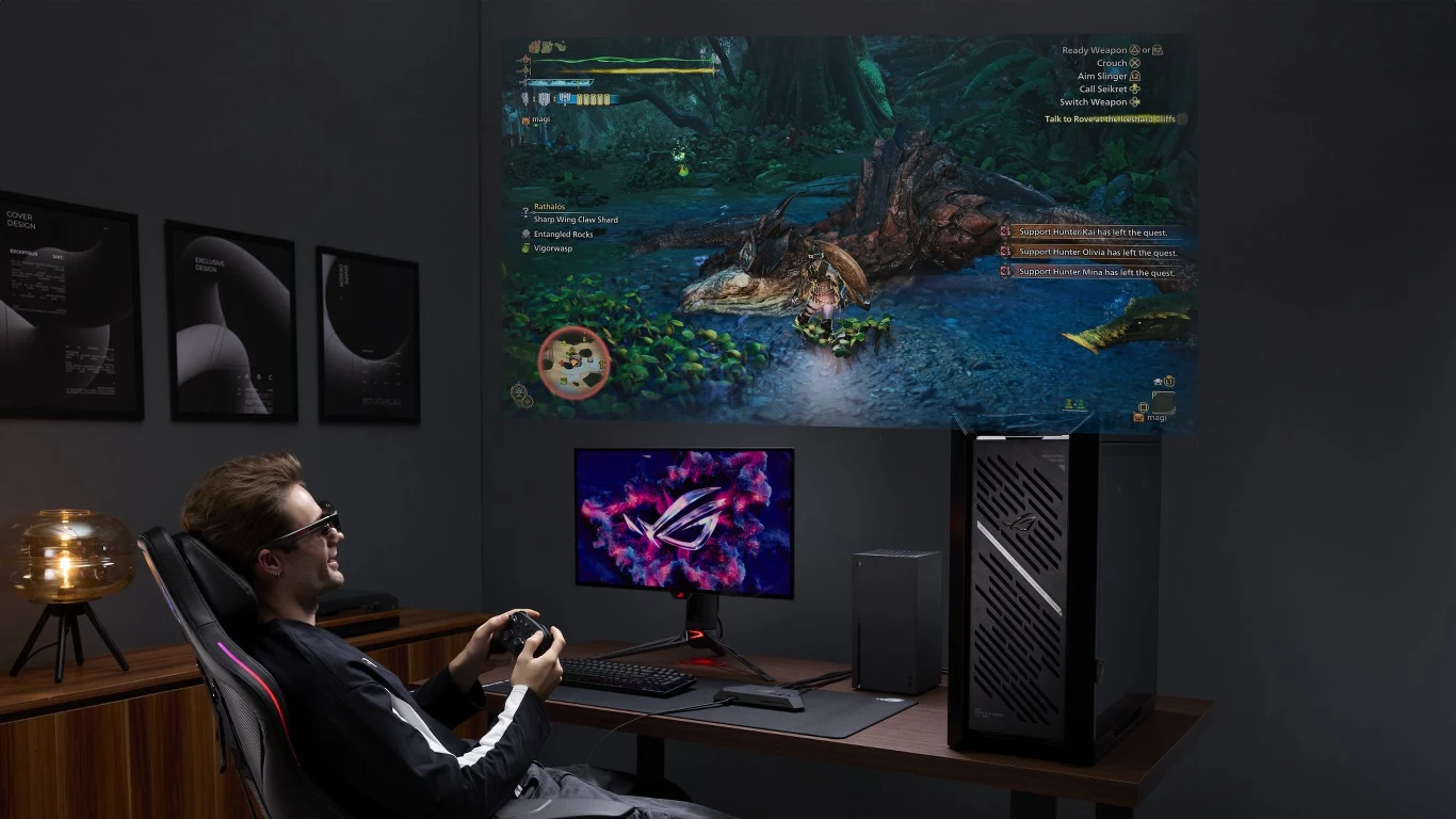 Gracz z padem w rękach siedzi przy biurku z nowoczesnym komputerem i monitorem, a na ścianie wyświetlana jest duża projekcja gry wideo z dynamiczną sceną walki z potworem.
