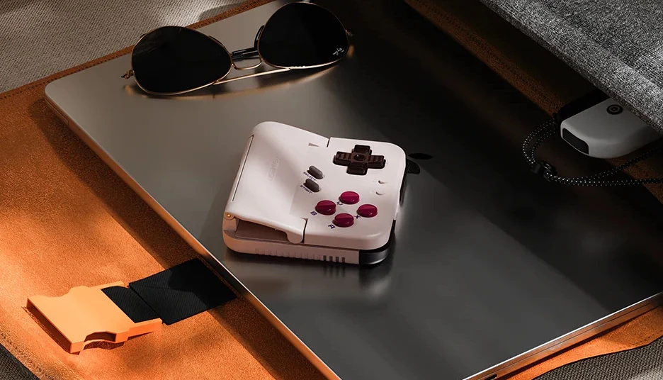 GameSir Pocket Taco Konsola przenośna w białym kolorze z ciemnymi przyciskami leży na zamkniętym laptopie, obok znajdują się okulary przeciwsłoneczne oraz pomarańczowy zatrzask paska torby.