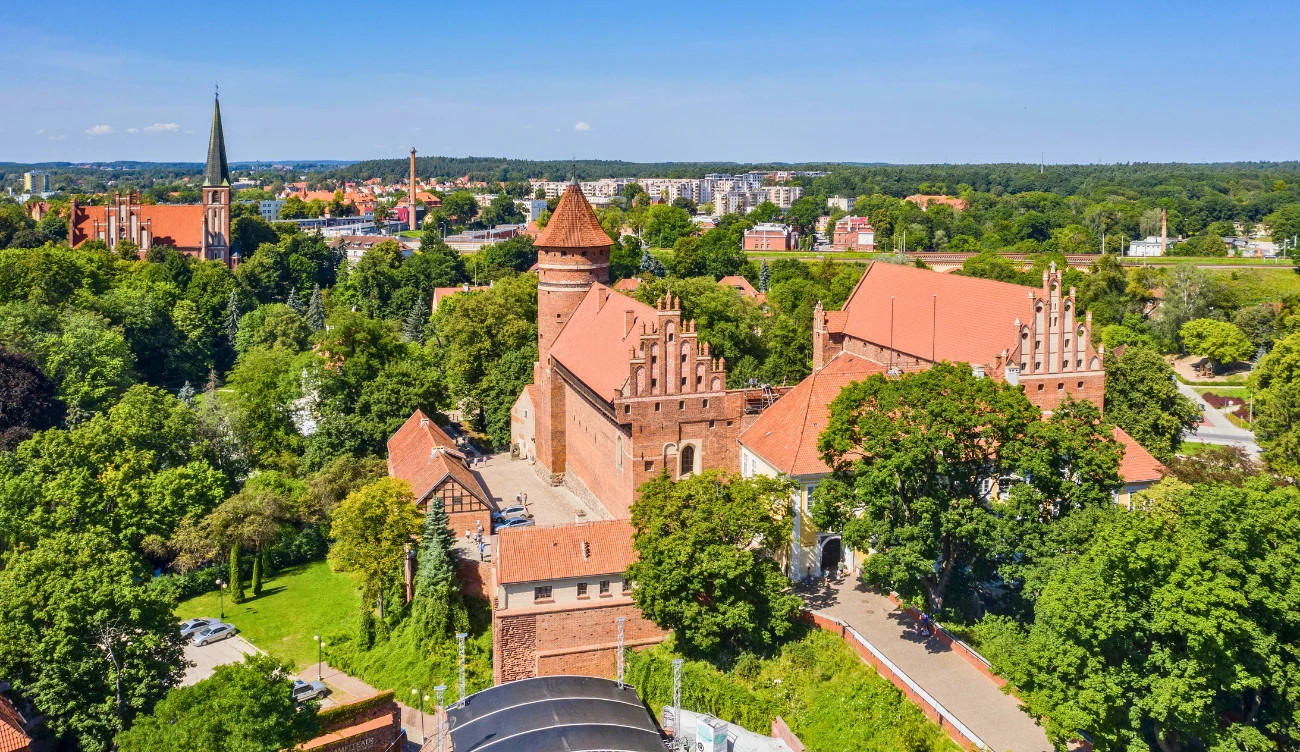 Ceglany zamek otoczony bujną zielenią i miejską zabudową na dalszym planie.