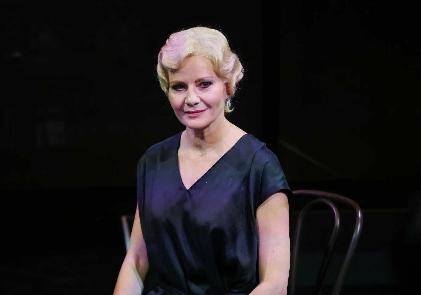 Małgorzata Kożuchowska na próbach do "Moralności pani Dulskiej" Kobieta z blond falowanymi włosami w eleganckiej, ciemnej sukni siedzi na krześle na tle ciemnej sceny.