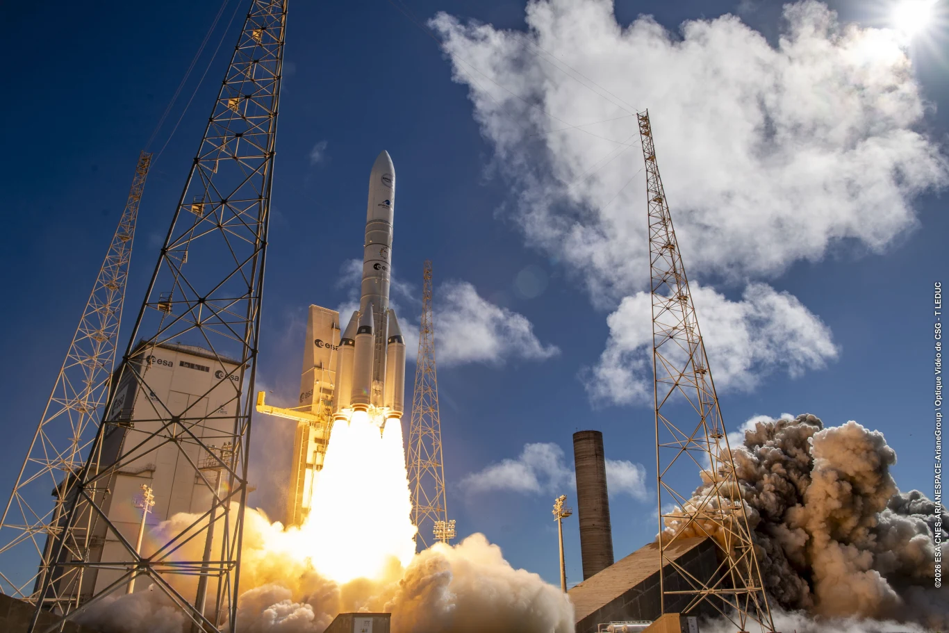 Ariane 64 Block 3 nie powstanie. Najpotężniejsza rakieta ESA zawieszona. Rakieta Ariane 64 w trakcie startu, widoczny jasny płomień i dym, wokół wieże zabezpieczające, niebo błękitne.