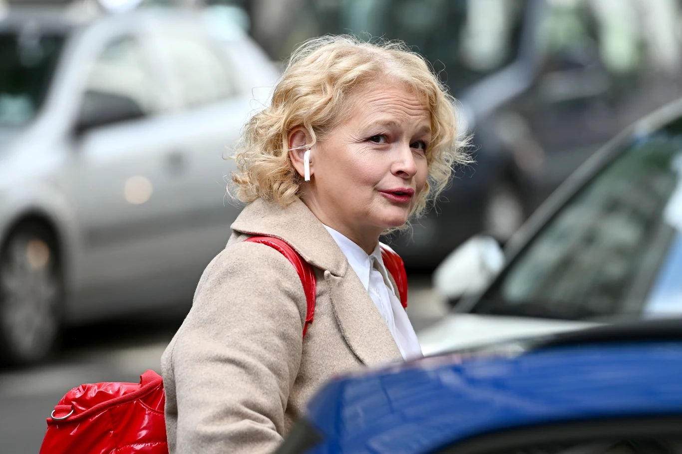 Dominika Ostałowska Dominika Ostałowska w beżowym płaszczu i z czerwonym plecakiem stoi na ulicy obok niebieskiego samochodu.