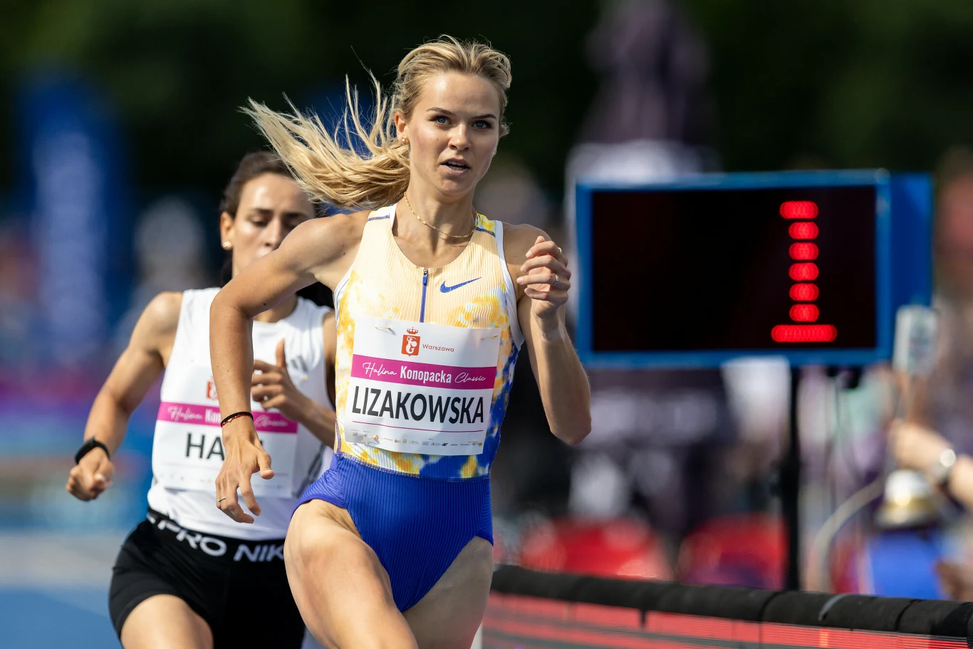 Weronika Lizakowska Dwie zawodniczki biegnące na lekkoatletycznej bieżni podczas zawodów, z przodu sportsmenka w żółto-niebieskim stroju z numerem startowym i wyraźnie widocznym intensywnym wysiłkiem, obok elektroniczny wyświetlacz z cyfrą jeden.