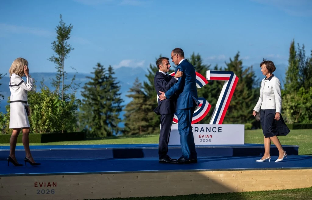 Dwóch mężczyzn w eleganckich garniturach obejmuje się na podwyższonej scenie z napisem 'FRANCE G7 ÉVIAN 2026', dwie kobiety w jasnych strojach zbliżają się do nich z obu stron, w tle widoczne drzewa i niebo.