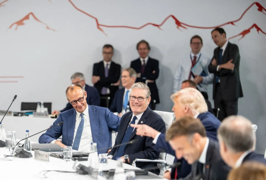 Friedrich Merz obok Keira Starmera na szczycie G7 Grupa elegancko ubranych mężczyzn siedzących przy stole konferencyjnym, uśmiechnięci i rozmawiający ze sobą w trakcie spotkania na wysokim szczeblu, z jasną ścianą ozdobioną liniowym motywem gór w tle.