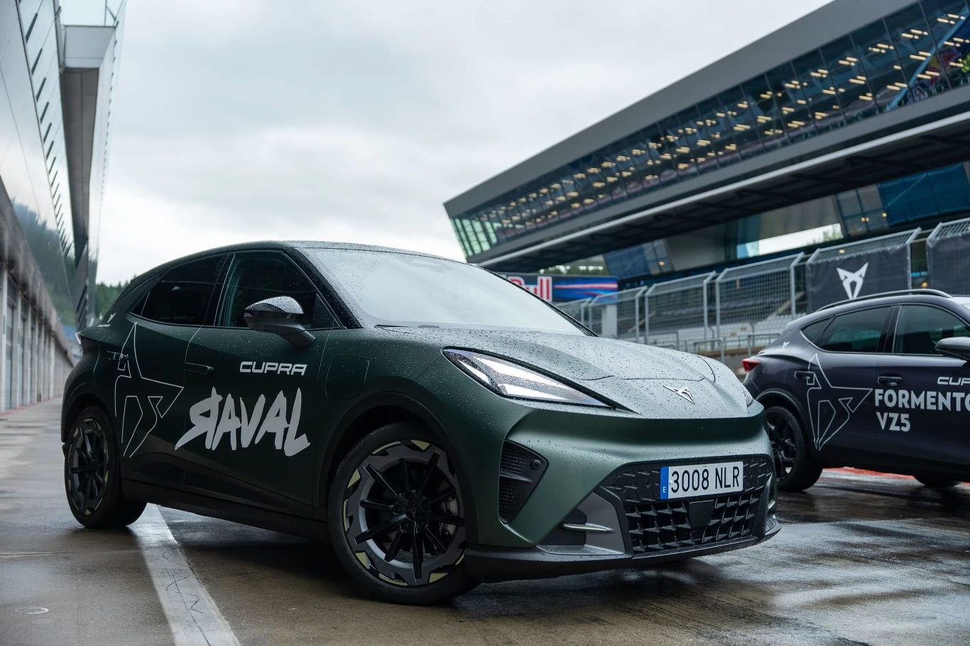 Nowoczesny, sportowy samochód Cupra Raval w ciemnozielonym kolorze z wyrazistym napisem na drzwiach, zaparkowany na torze wyścigowym obok innego auta tej samej marki.