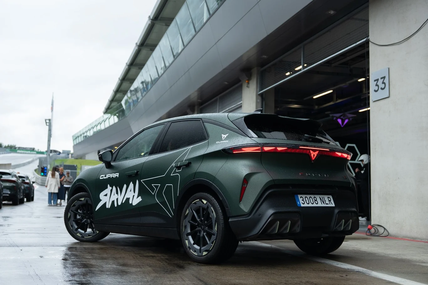 Nowoczesny SUV Cupra Tavascan z ciemnozielonym lakierem, wyrazistymi napisami na drzwiach i charakterystycznym, czerwonym światłem z tyłu, ustawiony przy wejściu do boksu na torze wyścigowym.