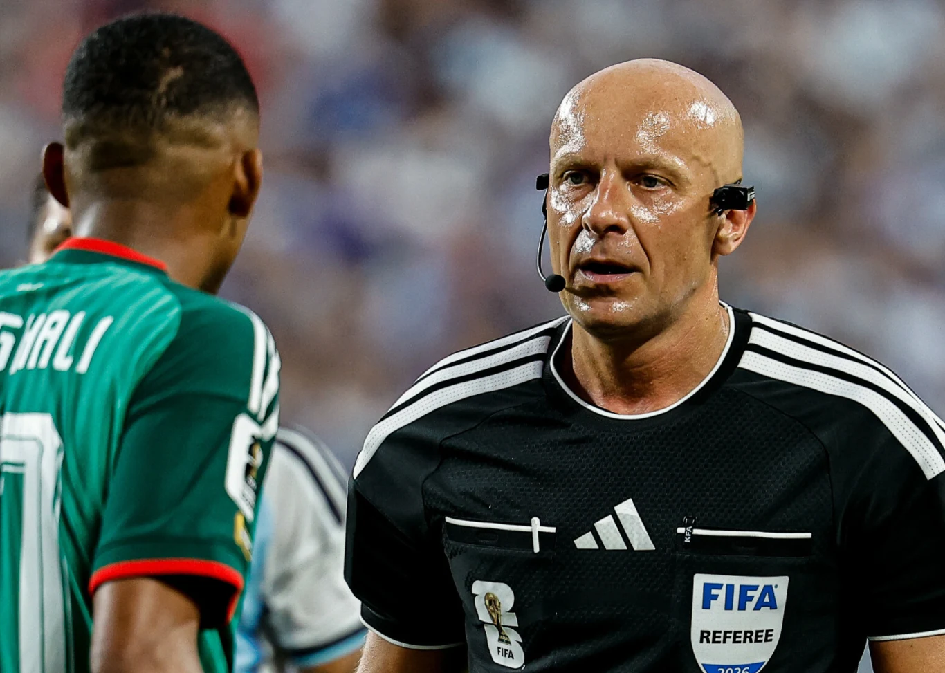 Sędzia piłkarski w czarnym stroju FIFA podczas rozmowy z zawodnikiem drużyny w zielonej koszulce na boisku podczas meczu.