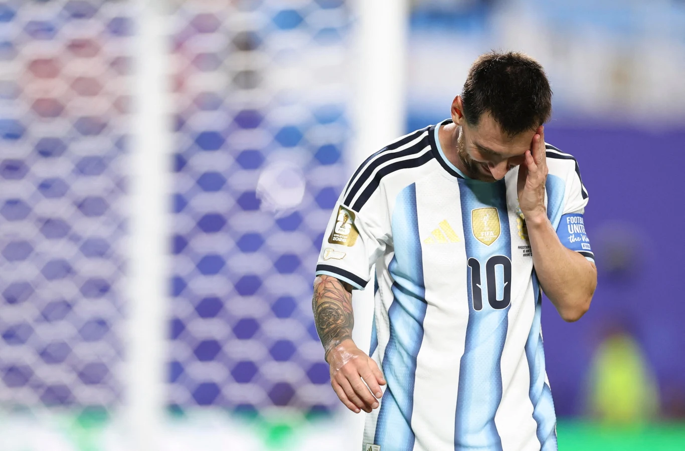 Lionel Messi Lionel Messi w biało-błękitnej koszulce Argentyny z numerem 10, z opuszczoną głową, dotyka twarzy ręką, w tle siatka bramki.