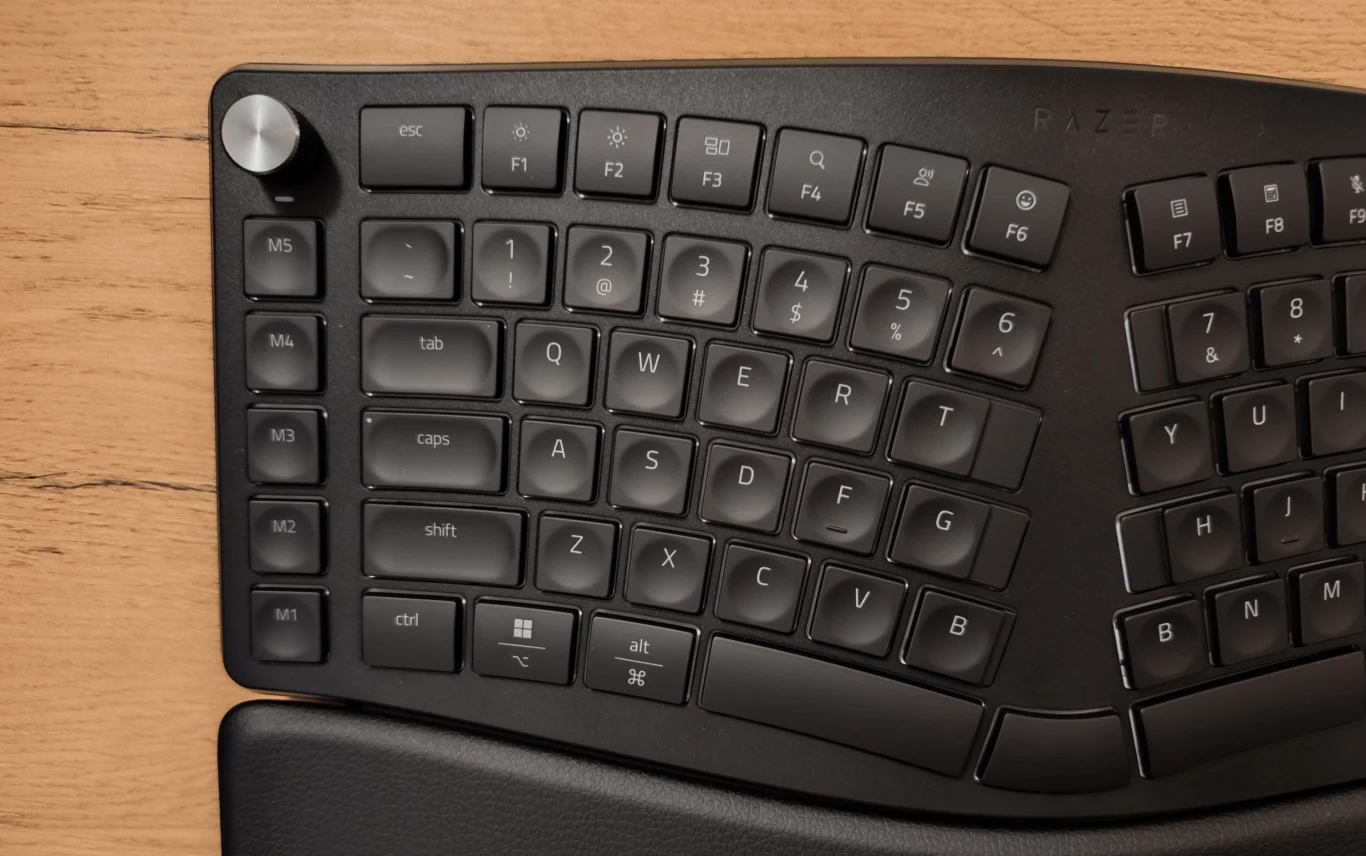 Czarna klawiatura ergonomiczna z podziałem na sekcje, dużym pokrętłem w lewym górnym rogu oraz wyraźnie wyprofilowanymi klawiszami na drewnianym biurku.