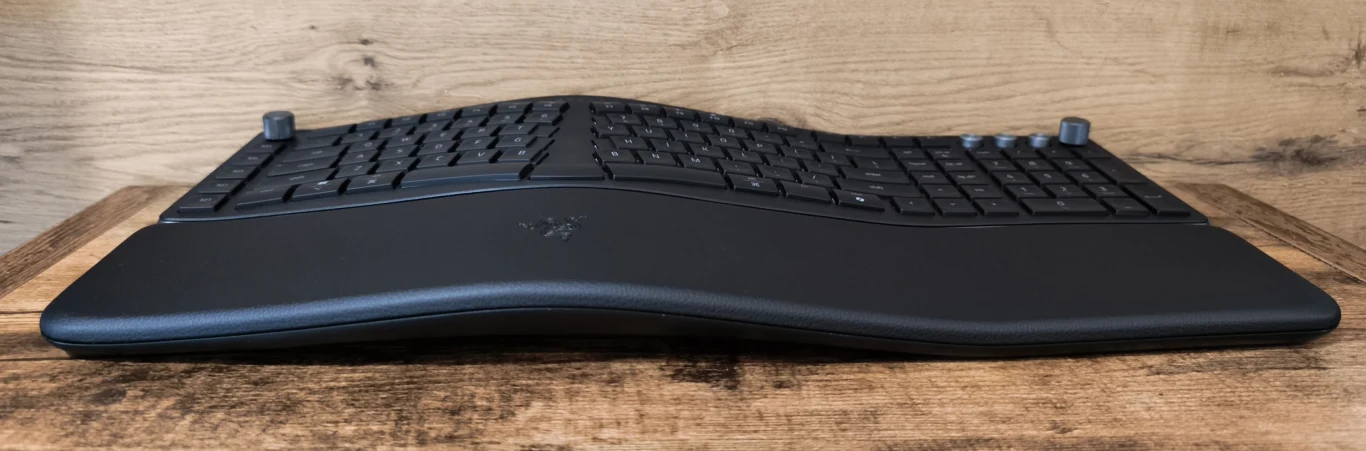 Czarna, ergonomiczna klawiatura z wyraźnie falistym profilem, rozdzieloną przestrzenią klawiszy i dużą podpórką na nadgarstki, ustawiona na drewnianej powierzchni na tle jasnej ściany.