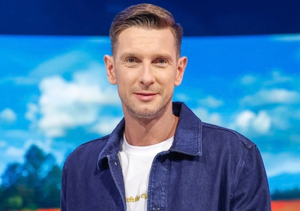 Łukasz Kadziewicz Łukasz Kadziewicz w jasnej koszulce i granatowej kurtce jeansowej na krajobrazu w studiu telewizyjnym.