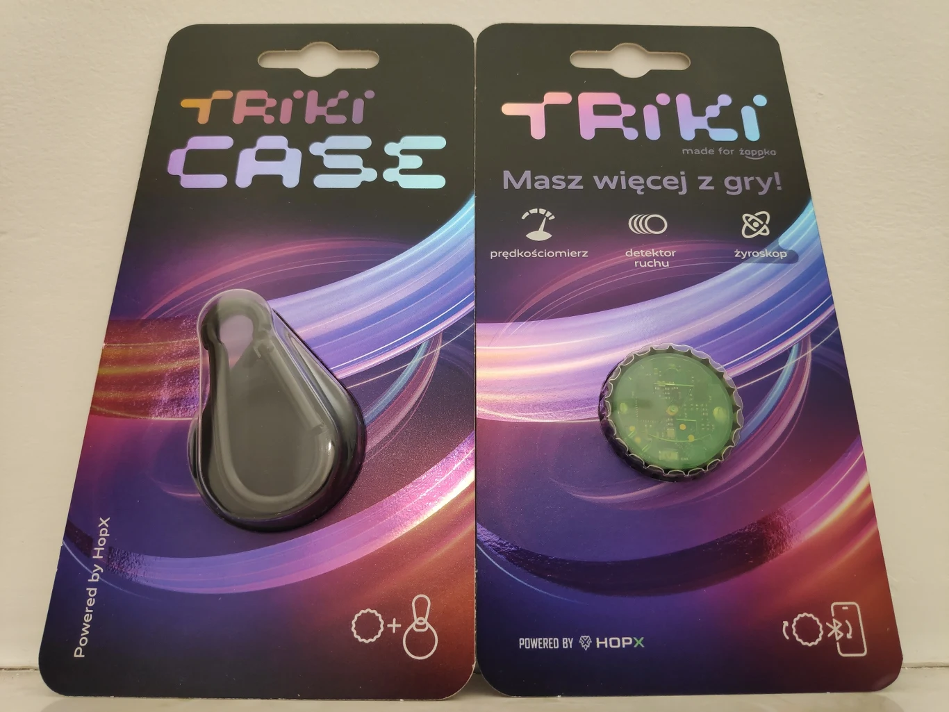Dwa opakowania produktów marki Triki, z których jedno zawiera czarne, przypominające kroplę etui, a drugie kapsel z zielonym nadrukiem, oba na tle kolorowej, dynamicznej grafiki z logo i ikonami funkcji.