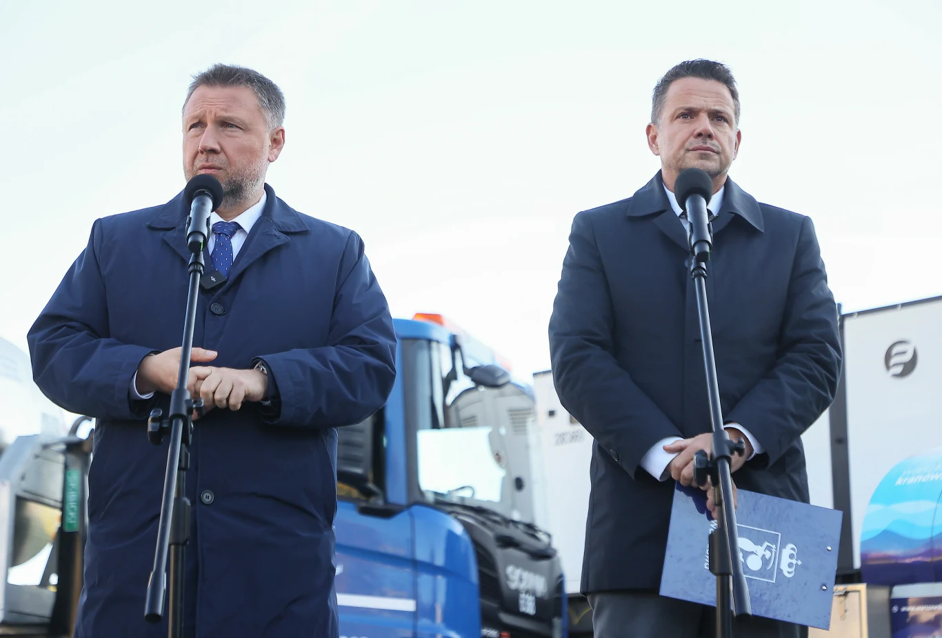 Dwaj mężczyźni w garniturach i płaszczach stoją przed mikrofonami, w tle widać samochody ciężarowe.