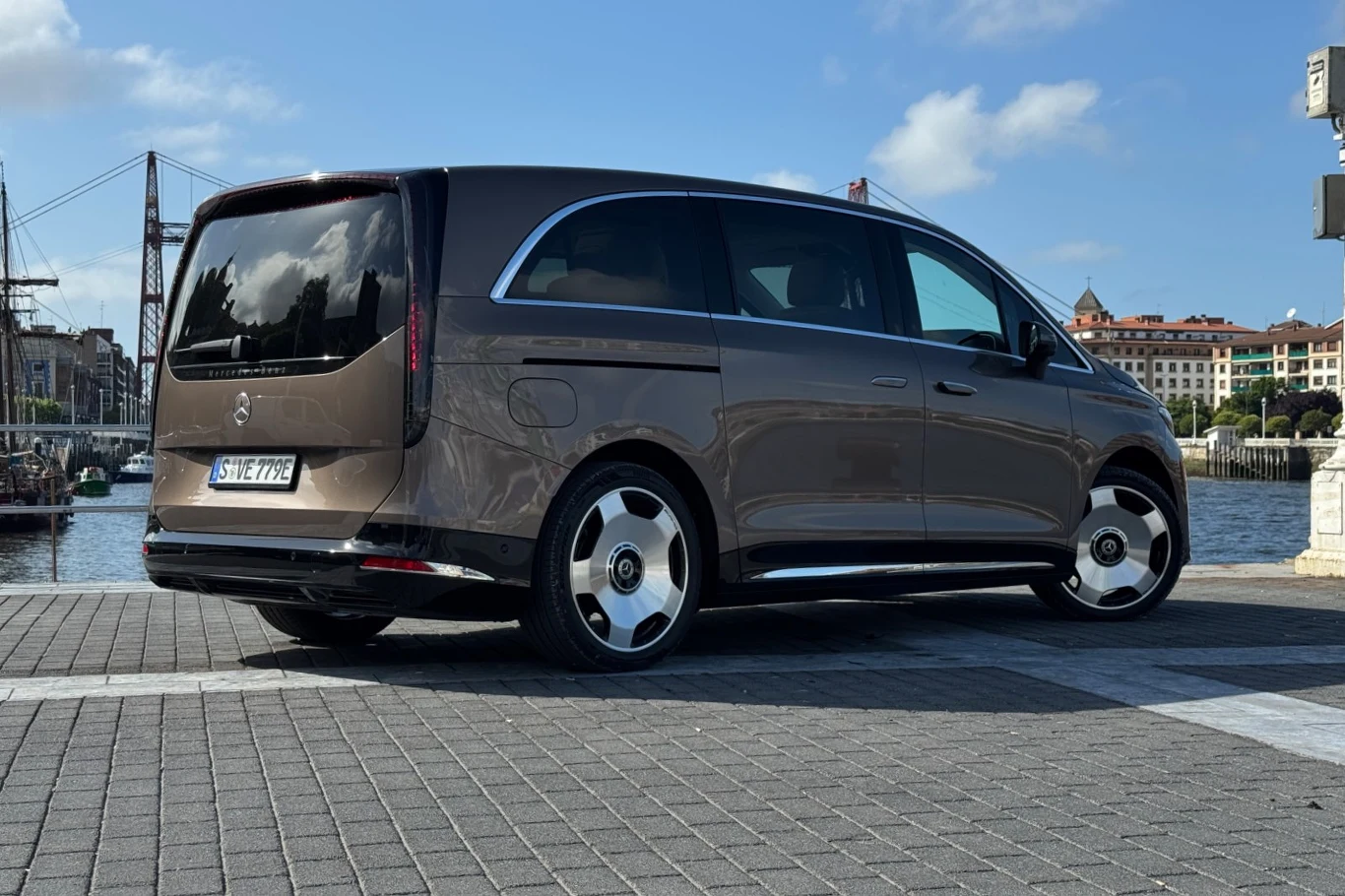 Brązowy minivan Mercedes-Benz zaparkowany na kostce brukowej nad wodą, z mostem kratownicowym i miejską zabudową w tle.