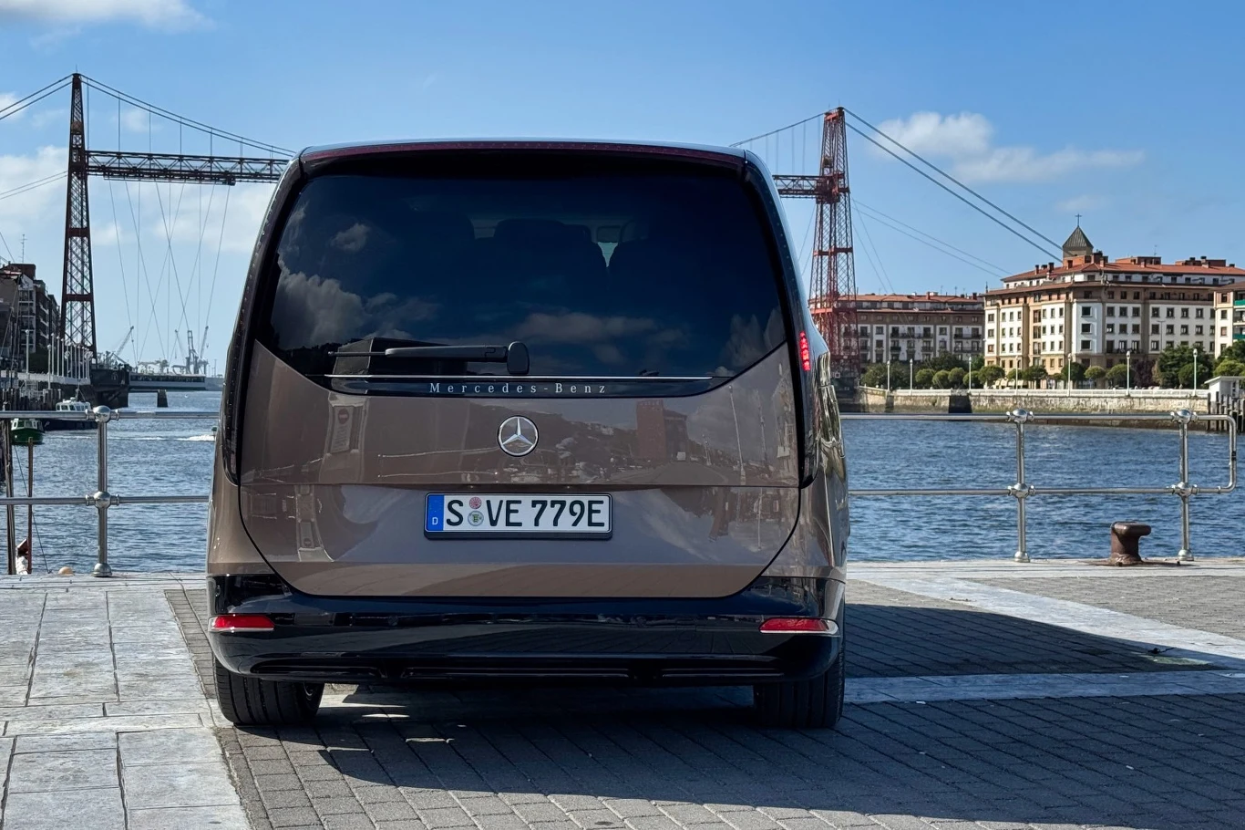 Luksusowy van Mercedes-Benz ustawiony tyłem przy nabrzeżu, w tle most wiszący i zabudowa miejska po drugiej stronie rzeki.
