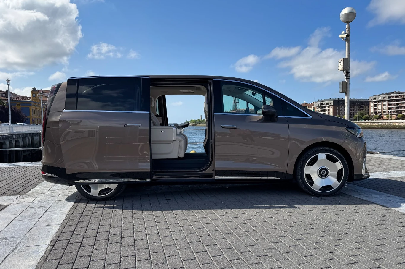 Brązowy nowoczesny minivan z otwartymi przesuwanymi drzwiami, zaparkowany przy nabrzeżu z widokiem na wodę i miejską zabudowę w tle.