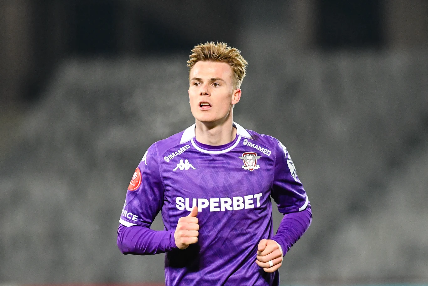 Tobias Christensen Piłkarz w fioletowej koszulce z logo sponsora i klubu, w trakcie biegu na boisku podczas meczu.