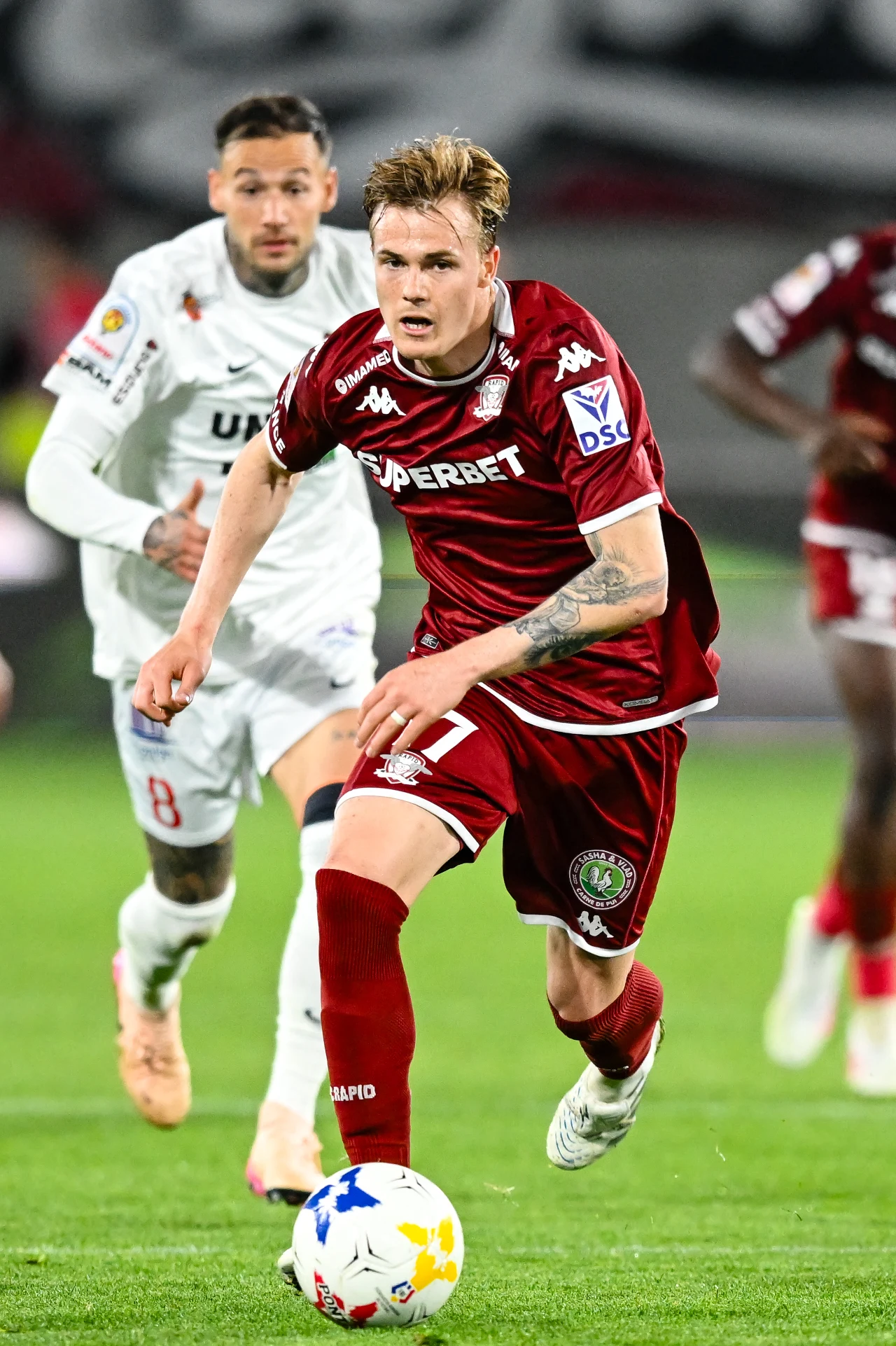 Tobias Christensen Piłkarz w bordowym stroju prowadzi piłkę podczas dynamicznej akcji meczowej, a tuż za nim biegnie rywal w białym stroju.